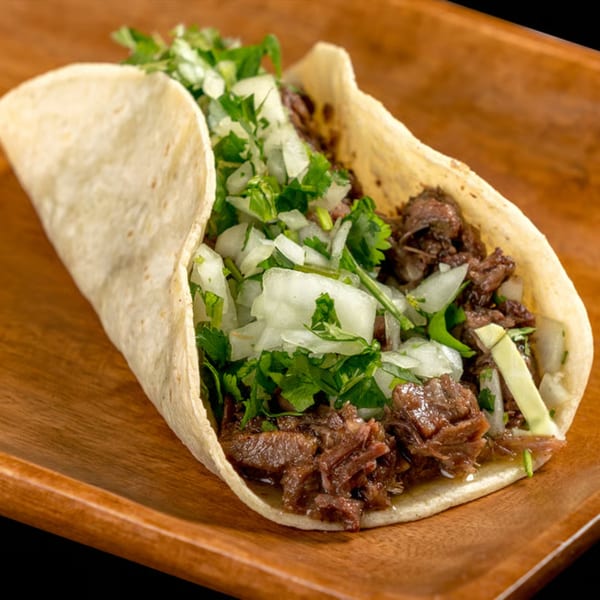 Close-up of a single taco filled with shredded meat, diced onions, and cilantro on a wooden plate.