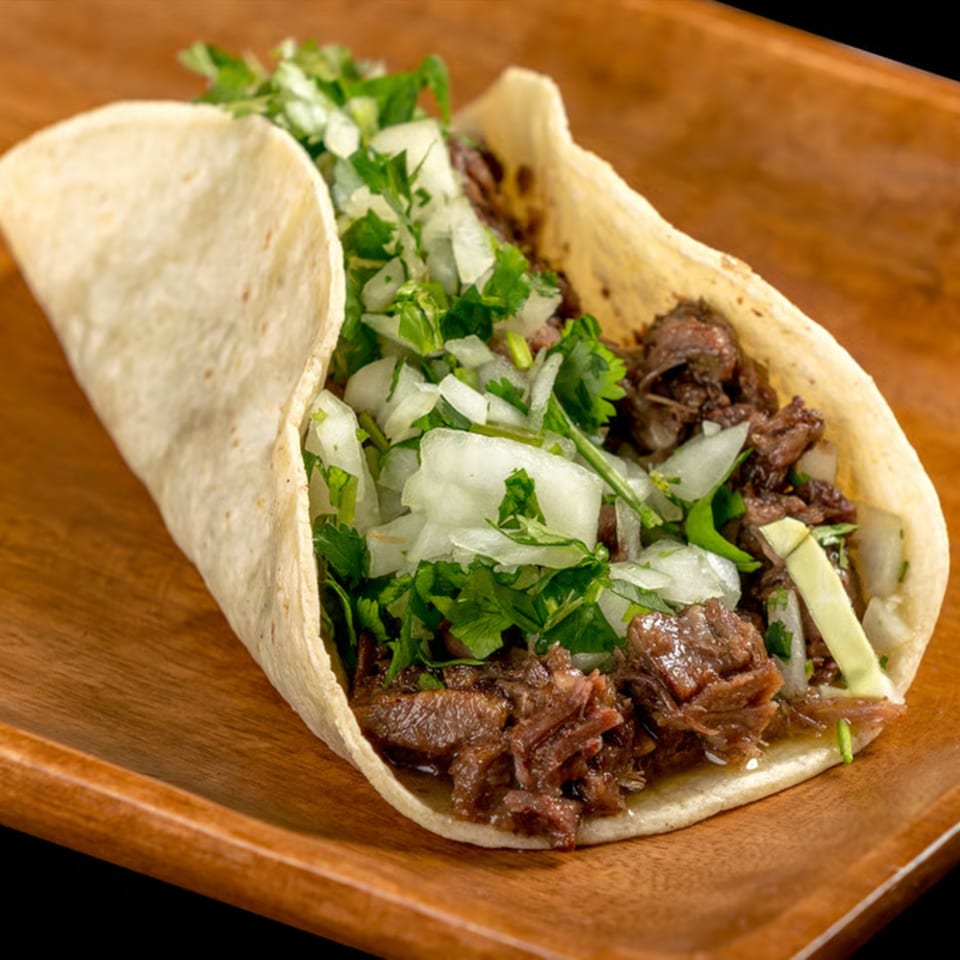 Close-up of a single taco filled with shredded meat, diced onions, and cilantro on a wooden plate.