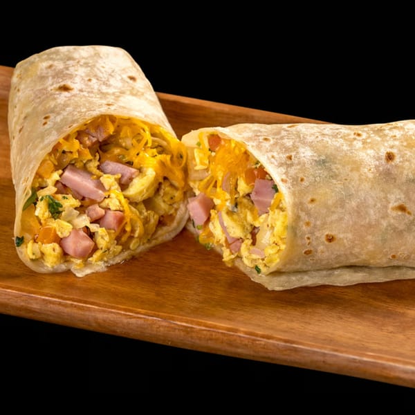 Breakfast burrito, cut in half, filled with scrambled eggs, ham, cheese, and vegetables on a wooden board.