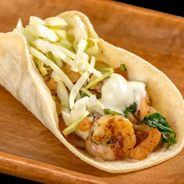 A shrimp taco with shredded cabbage, cilantro, and white sauce on a wooden tray.